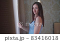 Young woman stands near the window in the bedroom, smiles and enjoys sunrise. She is dressed in 83416018