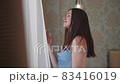 Young woman is standing near the window, stroking long dark hair with hands and enjoying sunrise 83416019