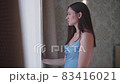 Young woman stands near the window in the bedroom and enjoys the sunrise. She is dressed in shirt of 83416021