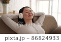 Young asian woman relaxing on comfortable sofa with eyes closed wearing headphones. Pretty mixed race lady enjoys listening chill music audio sound meditating feeling no stress at home. 86824933