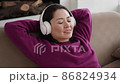 Young ethnic woman chilling on comfortable sofa with closed eyes wearing wireless headphones, asian female enjoys listening chill music and audio sound relaxing and meditating at home. 86824934