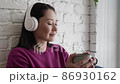 Attractive young woman holding cup of tea or coffee drinking warm beverage at home. Young asian girl in headphones listening chill music with morning drink looking through window enjoying and dreaming 86930162