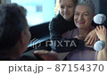 Mature white-haired woman checking eye wrinkles in front of mirror. Portrait of happy grandmother and teen granddaughter. Two generations of the same family 87154370