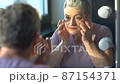 Under eye masks for puffiness, wrinkles, dark circles. Eye cream concept. Smiling senior gray haired woman in pink shirt, applying eye cream while looking in the mirror 87154371
