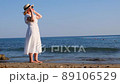 woman in a straw hat and white dress puts on sunglasses and walks along the seashore and admires the 89106529