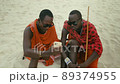 Medium Shot of Happy Maasai Tribe Men Sitting on the Sand in the Desert Watching Smartphone and Showing Thumb Up Sign. 89374955