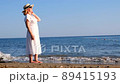 mature woman in sunglasses in a straw hat and white dress walks along the blue sea coast on a sunny 89415193