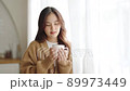 Young asian woman standing beside window and holding mug in living room at home 89973449
