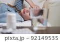 Tracking shot of a young woman lies sick on a couch with a headache or flu. There are tablets and a glass of water in front of her on the table. Blurred foreground. 92149535