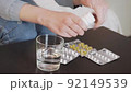 A young woman takes out a tablet from a bottle, takes a glass of water and takes medicine. Only hands and pills in frame. There are many tablets on the table. 92149539