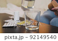 A young woman pours water into a glass before taking medicine. There are many pills on the table. 92149540