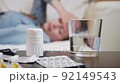 Tracking shot of a young woman lies sick on a couch with a headache or flu. There are tablets and a glass of water in front of her on the table. Blurred background. 92149543