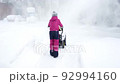 Child in crimson winter overalls and hat among snowdrifts during snowfall in yard of house removes snow with snowplow, back view. Lot of snow. Slow motion, 4k. 92994160