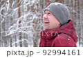 Laughing man in casual clothes looking up at the falling snow in a winter park or forest. Portrait of a young man with a friendly face enjoying the snowfall. Real people. Slow motion. Copy space. 92994161