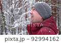 Smiling man in casual clothes looking up at the falling snow in a winter park or forest. Portrait of a young man with a friendly face enjoying the snowfall. Real people. Slow motion. Copy space. 92994162