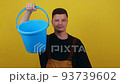 A young guy gardener or cleaner in uniform holds a blue bucket in his hands and points his finger at it 93739602