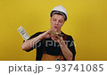 Foremans salary. Close-up photo of a happy young man in a suit and construction helmet, who is holding a wad of cash in his right hand and showing thumbs up. 93741085