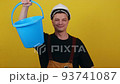 A young guy gardener or cleaner in uniform holds a blue bucket in his hands and points his finger at it 93741087