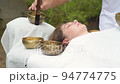 Young woman laying on a massage bed during a sound massage therapy with tibetan singing bowl outdoor Young woman laying on a massage bed during a sound massage therapy with tibetan singing bowl outdoor 94774775
