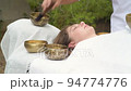 Young woman laying on a massage bed during a sound massage therapy with tibetan singing bowl outdoor Young woman laying on a massage bed during a sound massage therapy with tibetan singing bowl outdoor 94774776