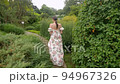 fashionable woman in a white floral dress walks around the estate with trimmed bushes and trees on the lawns in summer 94967326