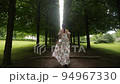 fashionable woman in a white dress walks up a grove of tall trimmed trees 94967330