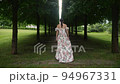fashionable woman in a white dress walks up a grove of tall trimmed trees 94967331