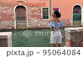 A young female tourist walks around Venice and wishes for photos on her mobile phone. A brunette woman admires the scenery of Venice at sunset. she smiles cheerfully in venice italy. her walk on the 95064084