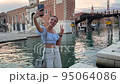 funny energetic woman makes video call using mobile internet on her smartphone. she is a tourist walking around Venice in Italy and posing against the backdrop of the Arsenal 95064086