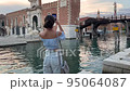 A young female tourist walks around Venice and wishes for photos on her mobile phone. A brunette woman admires the scenery of Venice at sunset. she smiles cheerfully in venice italy. her walk on the 95064087
