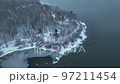 Fly Over snowy shore of unfrozen Lake with snow covered pines in foreground. Aerial view of a winter pine forest in snowfall. Top view of snow-covered pine trees. Beautiful winter forest landscape. 97211454
