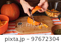 A woman slices an orange pumpkin with a knife into slices to serve on a wooden table. Conceptual autumn food in a cozy dark kitchen with yellow flowers 97672304