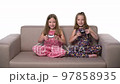 Two joyful pre-teen twin girls playing video game with joystick sitting on sofa 97858935