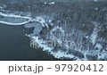 Fly Over snowy shore of unfrozen Lake with snow covered pines in foreground. Aerial view of a winter pine forest in snowfall. Top view of snow-covered pine trees. Beautiful winter forest landscape. 97920412