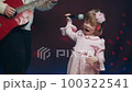 Little girl on stage in vintage dress, she sings into microphone and dances, her father plays acoustic guitar. Color music is shining. Performance on stage. Early development of children 100322541