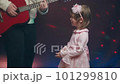 Little girl on stage in pink vintage dress, she dances, her father plays acoustic guitar. Color music is shining. Performance on stage. Early development of children 101299810