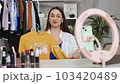 Caucasian influencer online trader seller woman female blogger sitting on couch recording video on phone camera on tripod sharing shopping experience showing sweater clothes. 103420489