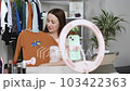 Young beautiful blogger mother sits front of mobile phone on tripod records fashion clothing video for social network online translation shows at camera enjoys clothes from online delivery 103422363