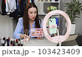 Young woman applying makeup on face with brush while recording vlog for social media via smartphone with tripod with LED ring lamp at table 103423409