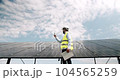 Male engineer speaking on smartphone on solar farm. Low angle man in uniform walking near photovoltaic panels and answering phone call against cloudy blue sky on sunny day on solar power station. 104565259