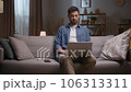 Man working late in evening. Male sitting at couch and using tablet. Thoughtfull man sitting at sofa and working distantly. Bearded man wearing wireless headphones. Man smiling at camera. 106313311