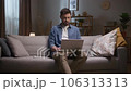 Man working late in evening. Male sitting at couch and using tablet. Thoughtfull man sitting at sofa and working distantly. Bearded man wearing wireless headphones. Man browsing at laptop. 106313313