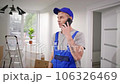 Man wearing blue uniform and cap. Builder in special overalls on repair object. Contractor talking by phone with foreman. Repairman making phone call. Man in uniform at background of ladder and boxes. 106326469