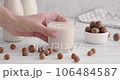 Hazelnut alternative milk. close-up video of hand taking a glass with vegan milk made of hazelnuts, grabbing a glass from white table, non-dairy drink, 4k horizontal video 106484587
