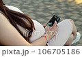 In a bustling public place, a pregnant young woman sits on a bench, deeply engrossed in examining an ultrasound photo of her baby on her smartphone. 106705186