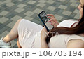 In a bustling public place, a pregnant young woman sits on a bench, deeply engrossed in examining an ultrasound photo of her baby on her smartphone. 106705194
