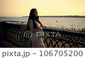 A soon-to-be mother chats on her phone while standing near the waterfront, tenderly touching her growing bump. 106705200