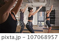 Team of multiethnic people doing group yoga in fitness center. People in sports uniforms performing exercises while standing on mats. Raising up their hands to relax body. Teamworking. 107068957