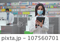 Female pharmacist holding tablet at foreground. Woman druggist working with gadget at workplace. Woman in white robe and mask near showcase with medicine. Pharmacists in drugstore. 107560701