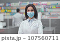 Close up view of female pharmacist with crossed arms. Portrait of woman pharmacist in drugstore. Woman wearing white robe and protective mask. Showcase with medicine. Caucasian druggist in pharmacy. 107560711